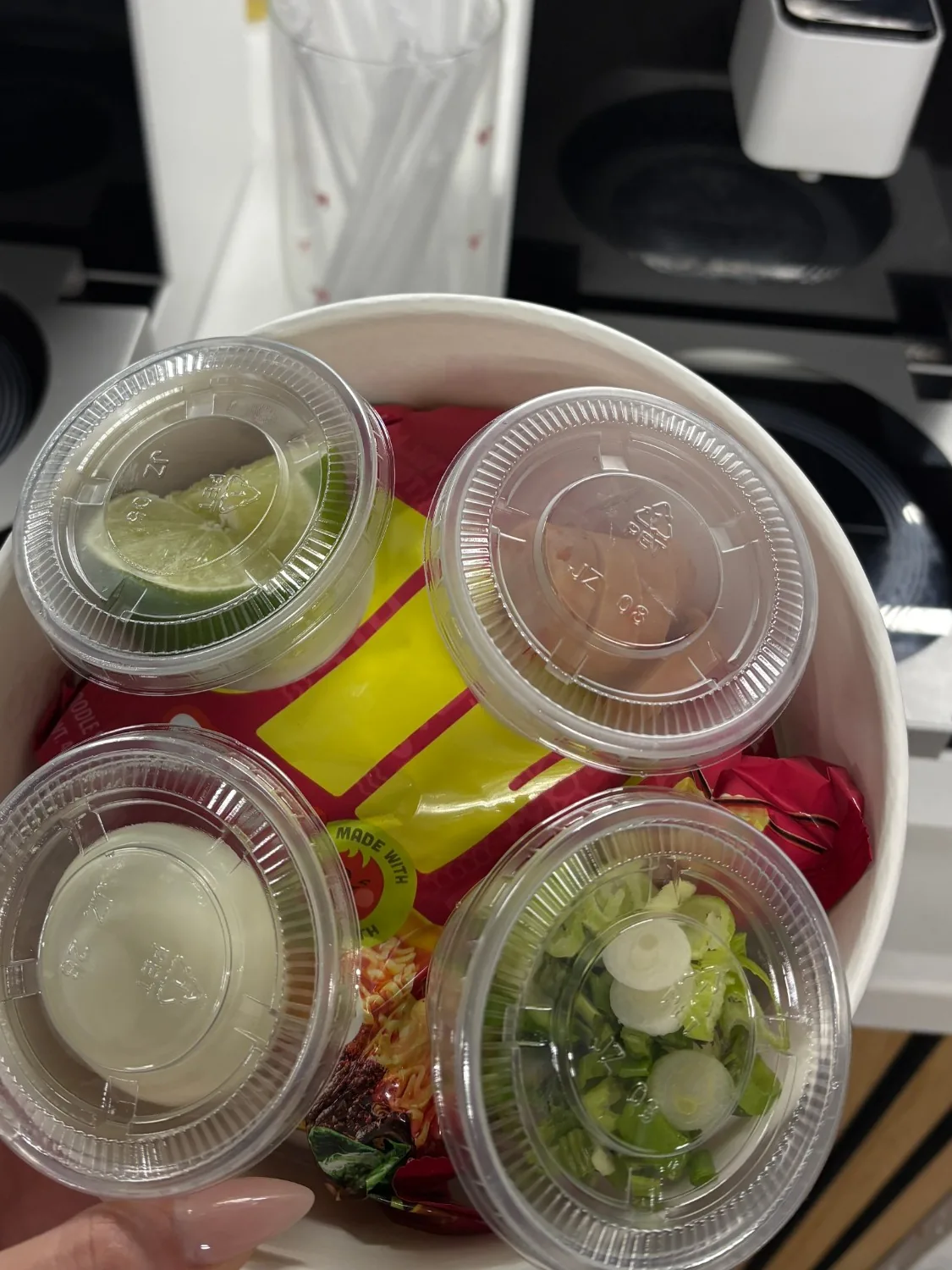 Four dollar-topping cups — lime, spicy sausage, a hard-boiled egg, and green onions — stacked on a ramen bowl
